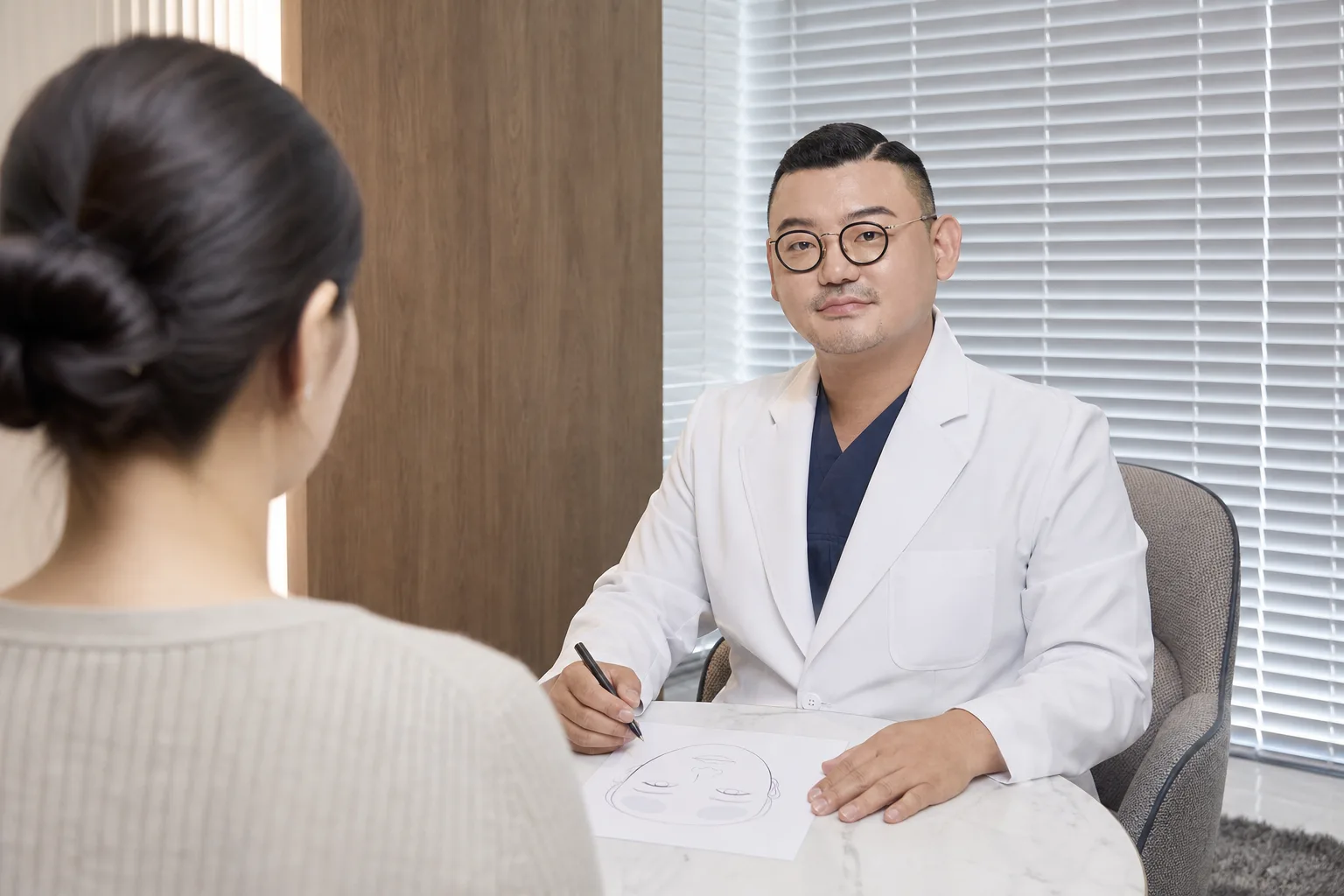Dr. Hongseok Kim sketching a face map with pen and paper while planning filler with a visiting patient, seen from behind, at VOS Dermatology, Gangnam
