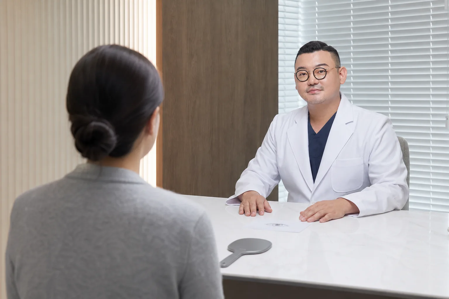 Dr. Hongseok Kim listening to a patient, seen from behind, across a desk with a hand mirror and an eye sketch during an under-eye consultation at VOS Dermatology, Gangnam